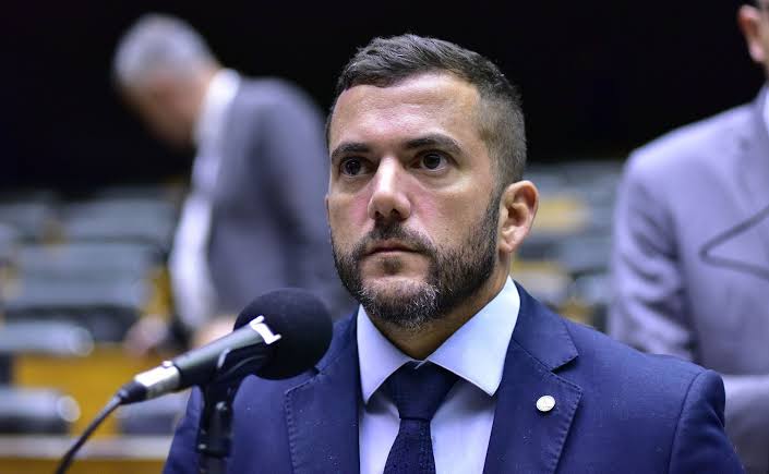 Deputado fluminense Carlos Jordy discursando com a bandeira do Brasil e o logotipo do Congresso Nacional ao fundo.